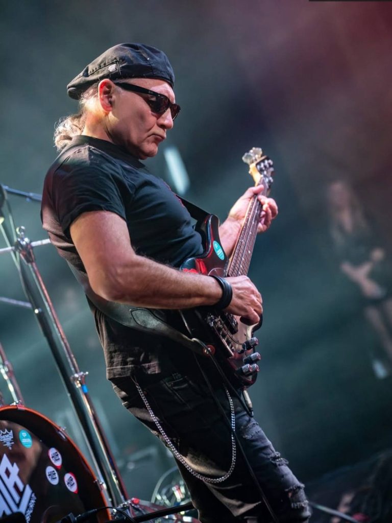 Basista z gitarą basową, w czapce i ciemnych okularach, w dymie scenicznym