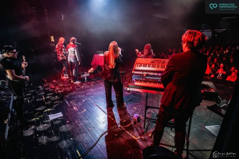 Koncert widziany z tyłu sceny: muzycy przy instrumentach w czerwonym świetle, przed nimi publiczność w sali
